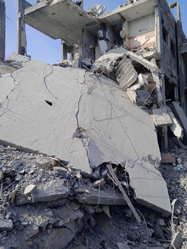 The image shows a heavily damaged multi-story building reduced to rubble. Large concrete slabs have collapsed, with exposed rebar and broken walls hanging precariously. Debris, including shattered bricks, twisted metal wires, and remnants of household items, is scattered across the ground. The destruction appears extensive, leaving only fragments of the original structure intact. The scene conveys the aftermath of intense conflict or bombing, highlighting the devastating impact on infrastructure.